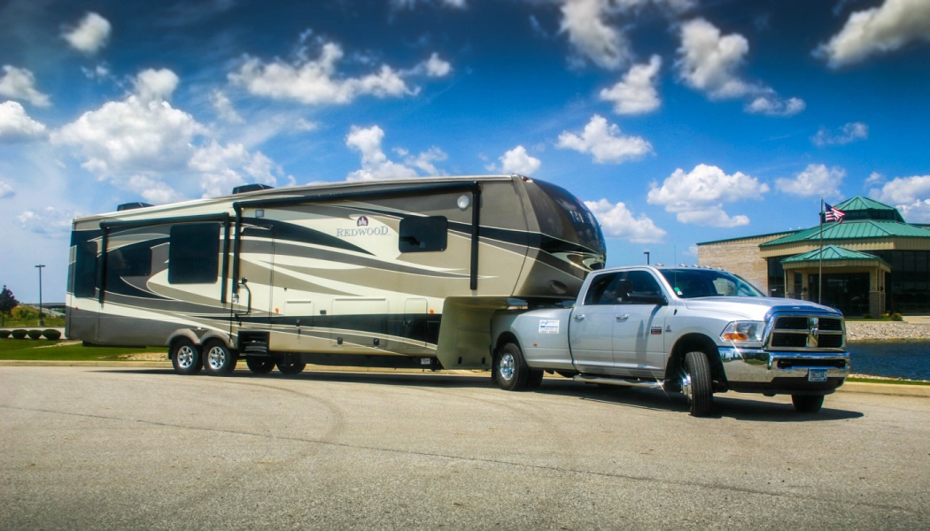 Silver pickup truck hitched to a large Redwood fifth-wheel RV trailer under a blue cloudy sky.