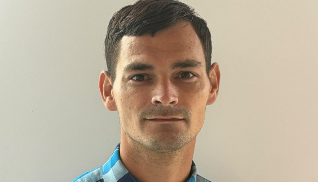 Close-up headshot of a man with short dark hair and light stubble, wearing a blue plaid shirt.