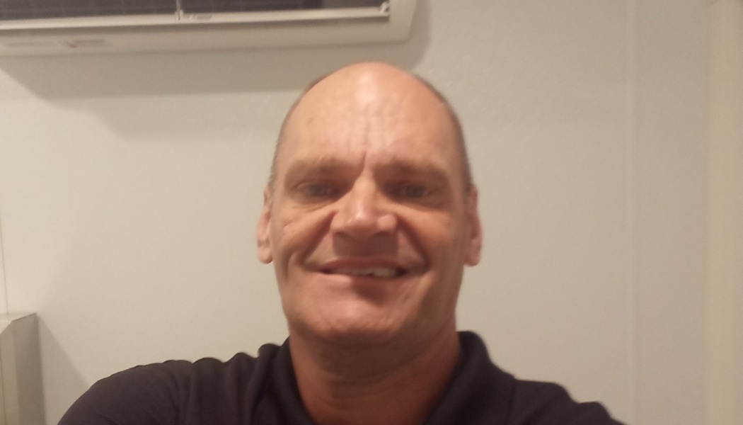 A close-up selfie of a smiling, bald man wearing a dark collared shirt.