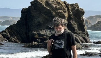 A bearded man looking down on a rocky beach with a large rock formation and ocean waves behind him.