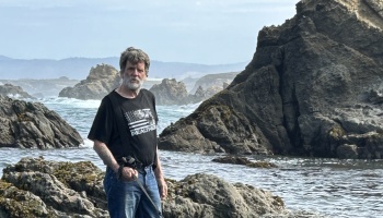 An older bearded man stands on a rocky ocean coastline surrounded by large dark cliffs and crashing waves.