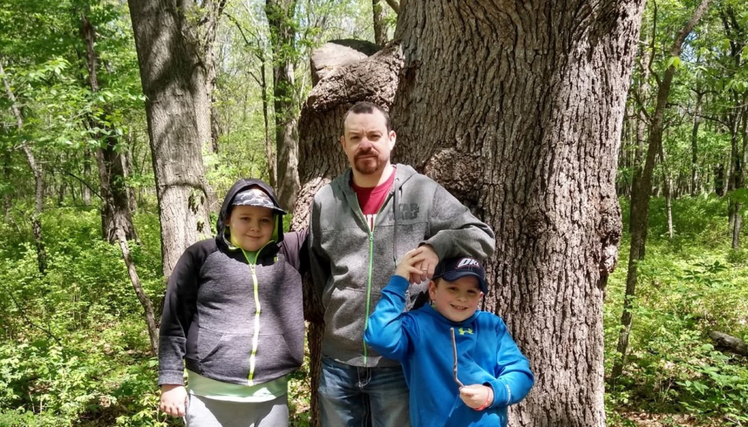 A father stands with his two young sons in front of a large tree in a green forest.
