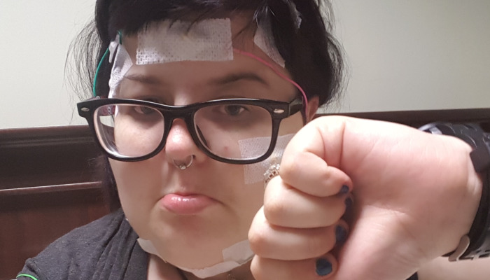 A person with glasses holds up a fist, wearing medical sensors taped to their forehead, cheek, and chin.