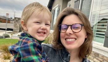 A smiling woman with glasses holds a happy young toddler wearing a colorful plaid jacket outside a house.