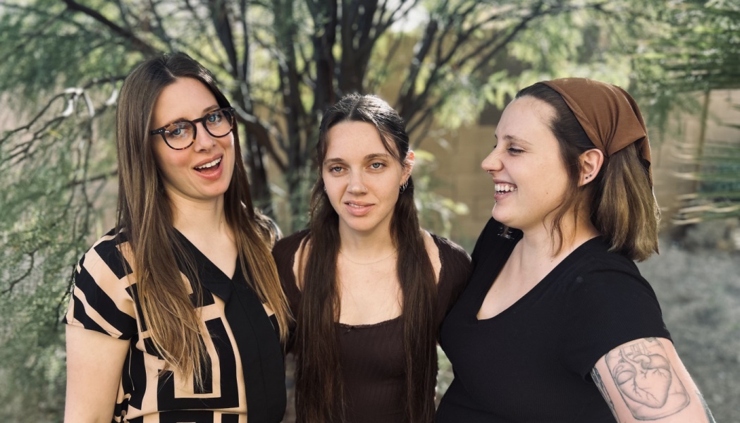 Three young women standing side-by-side outdoors, smiling and laughing in front of green trees.