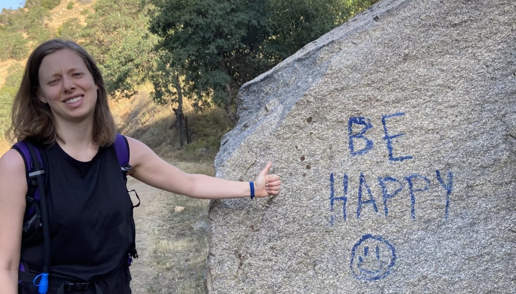 A smiling hiker gives a thumbs-up next to a large rock with blue writing that reads "BE HAPPY" and a smiley face.