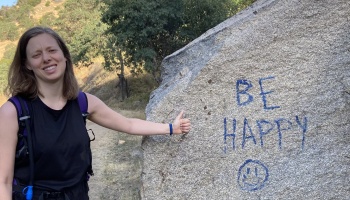 A smiling hiker gives a thumbs-up next to a large rock with blue writing that reads "BE HAPPY" and a smiley face.