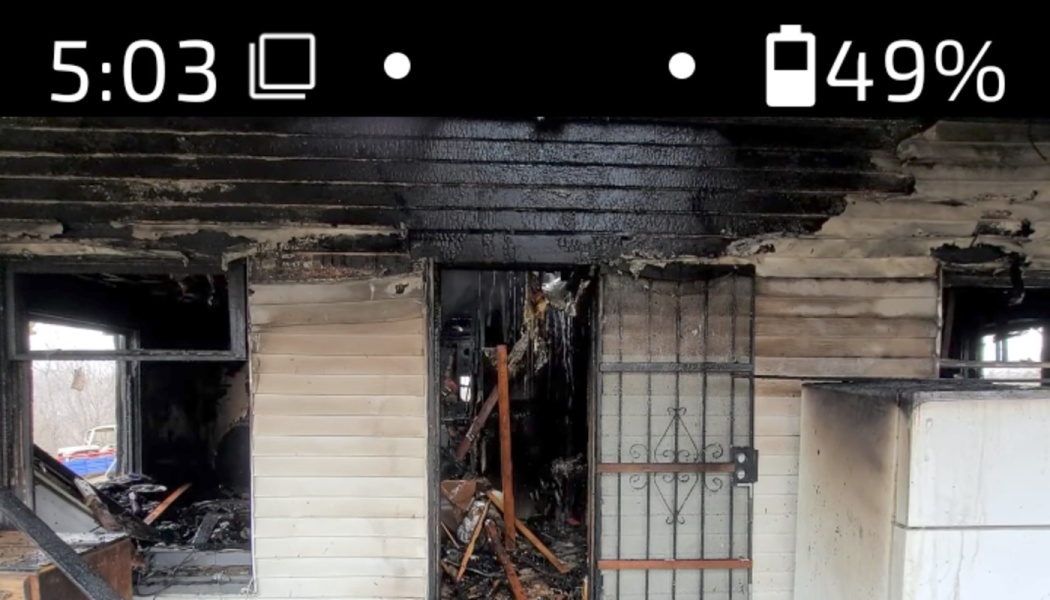 Severely fire-damaged home with charred siding, broken windows, and a gutted interior visible through the doorway.