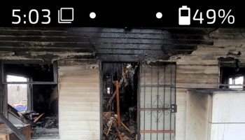 Severely fire-damaged home with charred siding, broken windows, and a gutted interior visible through the doorway.