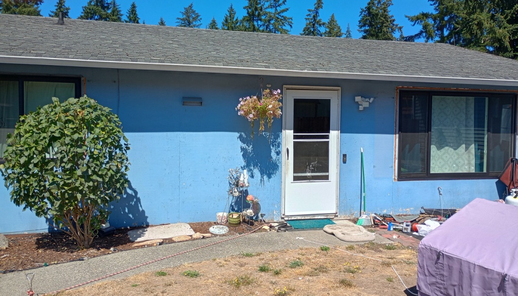 Exterior of a blue house featuring a white front door, large green bush, hanging flowers, and exposed window trim.