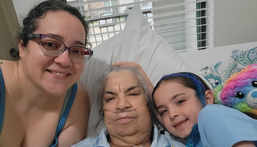 A woman, an elderly woman with an oxygen tube in bed, and a young girl smile together for a close selfie.