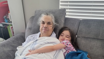 A young girl lovingly hugs an elderly woman wearing an oxygen cannula as they relax together on a couch.