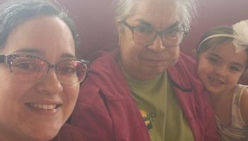 A woman with glasses smiling alongside an older woman and a young girl wearing a white bow.