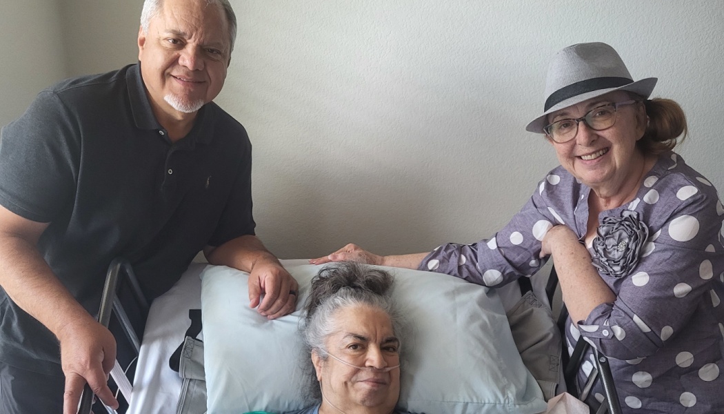 A smiling woman rests in a medical bed with a nasal cannula, flanked by a supportive man and woman.