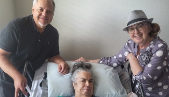 A smiling woman rests in a medical bed with a nasal cannula, flanked by a supportive man and woman.