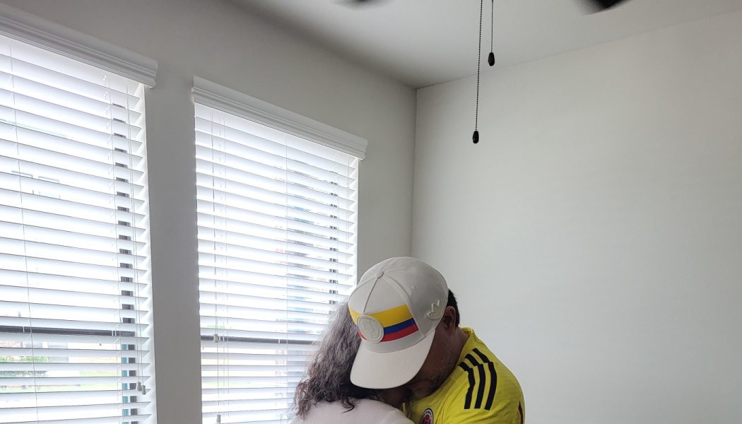 A man in a yellow jersey and Colombian flag hat lovingly embraces a woman near a window.