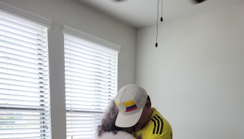 A man in a yellow jersey and Colombian flag hat lovingly embraces a woman near a window.
