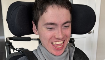 A young man with short dark hair smiles broadly while sitting in a wheelchair with head support pads.