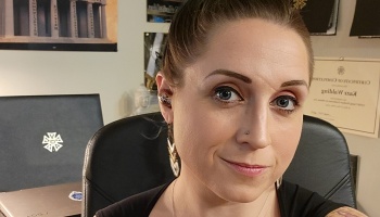 A woman with brown hair pulled back and blue eyes smiling softly at her office desk.
