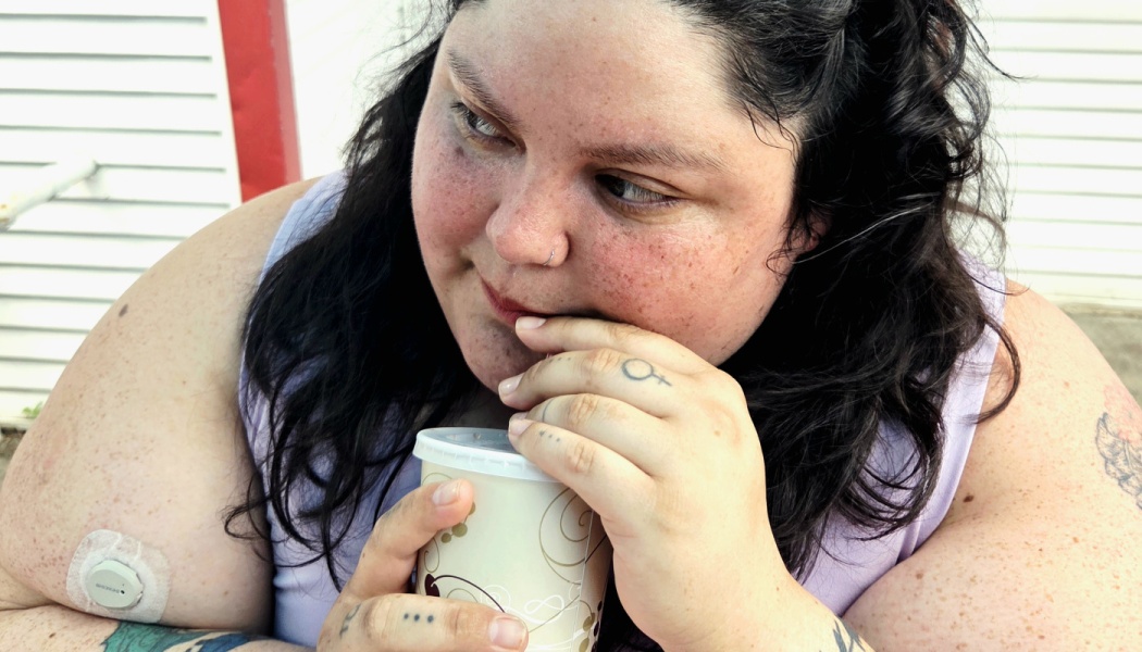 A freckled woman with dark wavy hair sips a drink through a straw while looking thoughtfully off to the side.