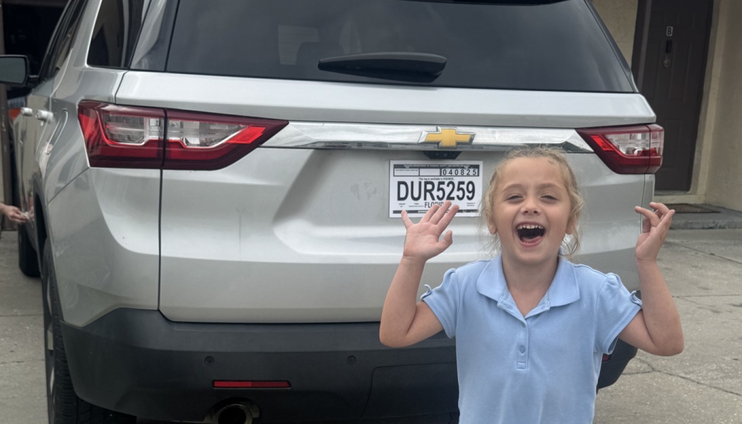 Excited young girl cheering with raised hands in front of a silver SUV.