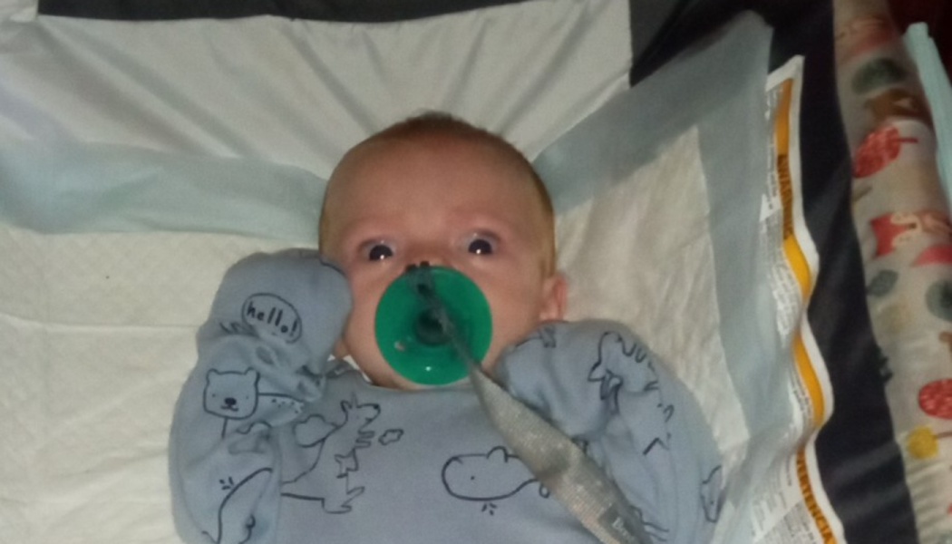 A wide-eyed baby in blue pajamas lies back with a green pacifier in their mouth.