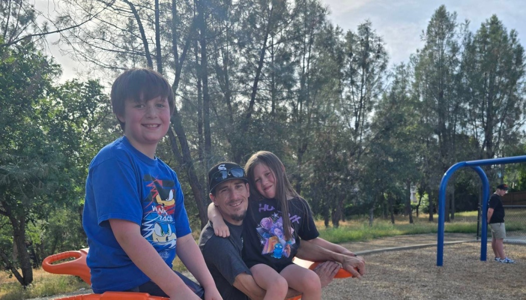 A smiling father sitting with his young son and daughter at an outdoor playground surrounded by trees.
