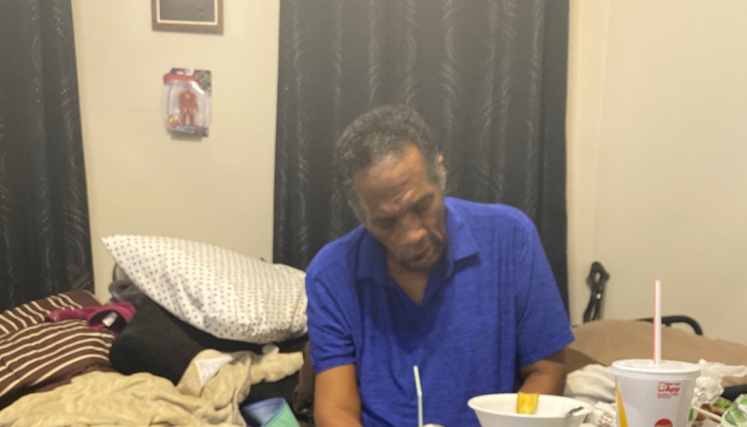 An older Black man in a blue polo shirt sitting indoors and looking down at a meal.