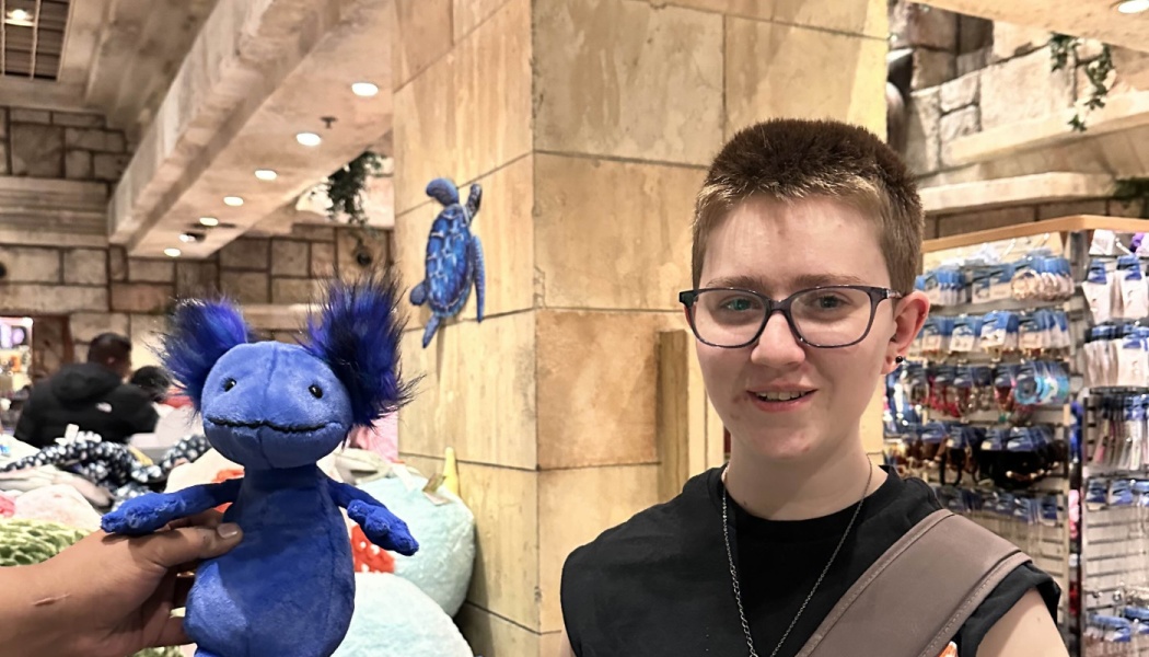 A smiling person with short hair and glasses stands in a gift shop next to a fluffy blue plush creature.
