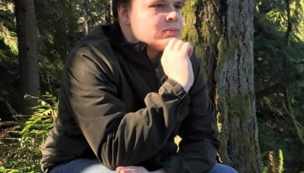 A young man sitting thoughtfully in a forest with his chin resting on his hand.