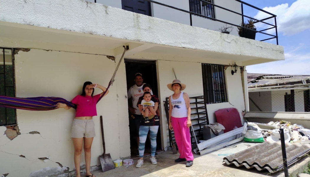 A family stands outside their damaged home with severe cracks on the exterior walls and debris scattered nearby.