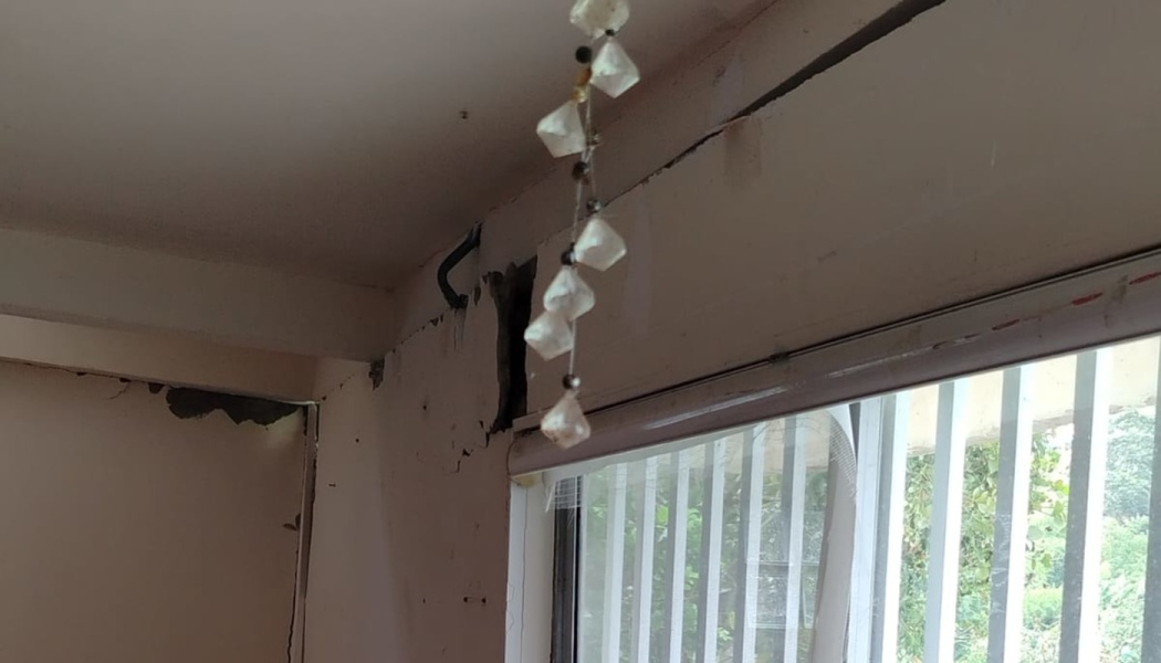 Severe cracks and broken plaster on an interior wall above a window with a hanging crystal decoration.