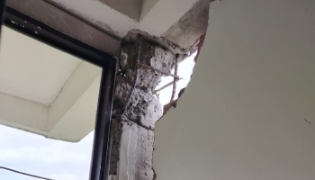 Severely damaged concrete column with exposed steel rebar and broken wall beside a window.