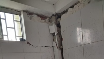 Severely damaged tiled wall with deep cracks and exposed concrete in the corner near a window.