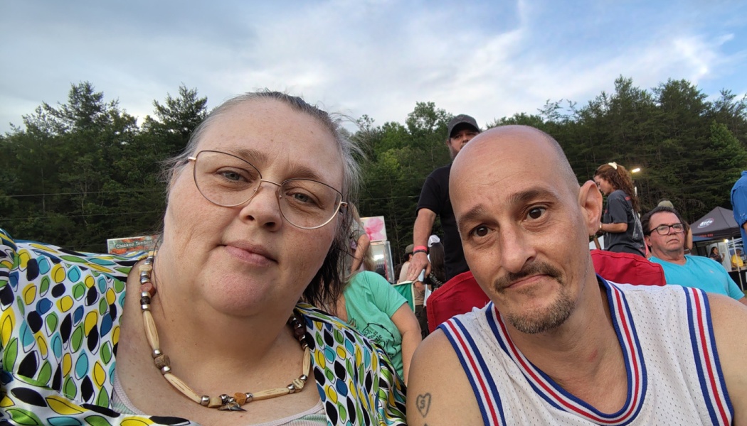 A woman with glasses and a bald man with a goatee taking a close-up selfie at an outdoor event.