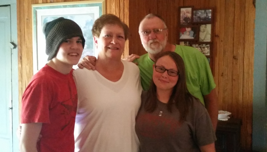 A smiling family of four poses closely together indoors in front of a wood-paneled wall.