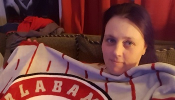 A woman sitting on a sofa, wrapped in a red and white Alabama fleece blanket.