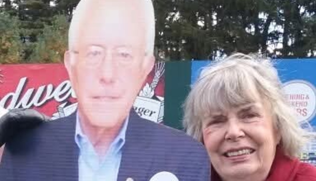 A smiling woman with gray hair poses beside a Bernie Sanders cardboard cutout.