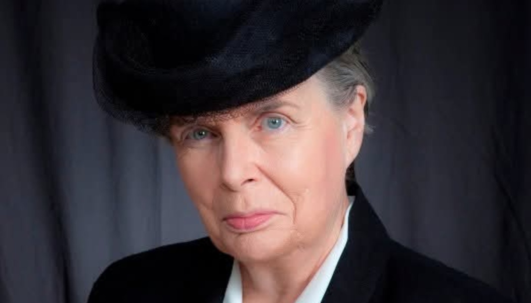 Close-up portrait of an older woman with blue eyes wearing a black vintage hat and suit jacket.