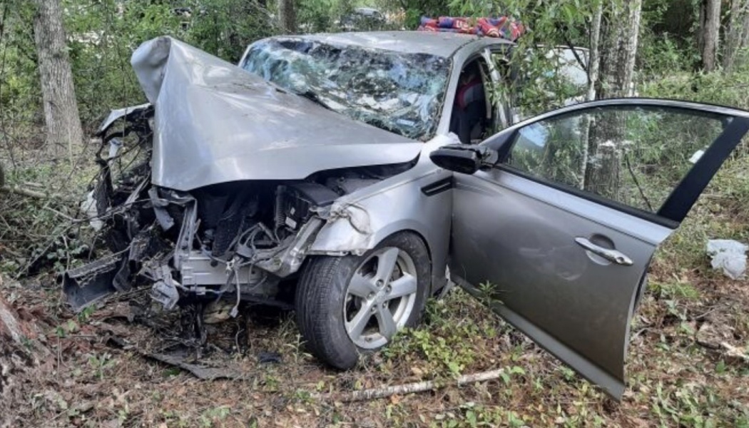 Severely damaged silver car with a heavily crumpled front end and open door in a wooded area.