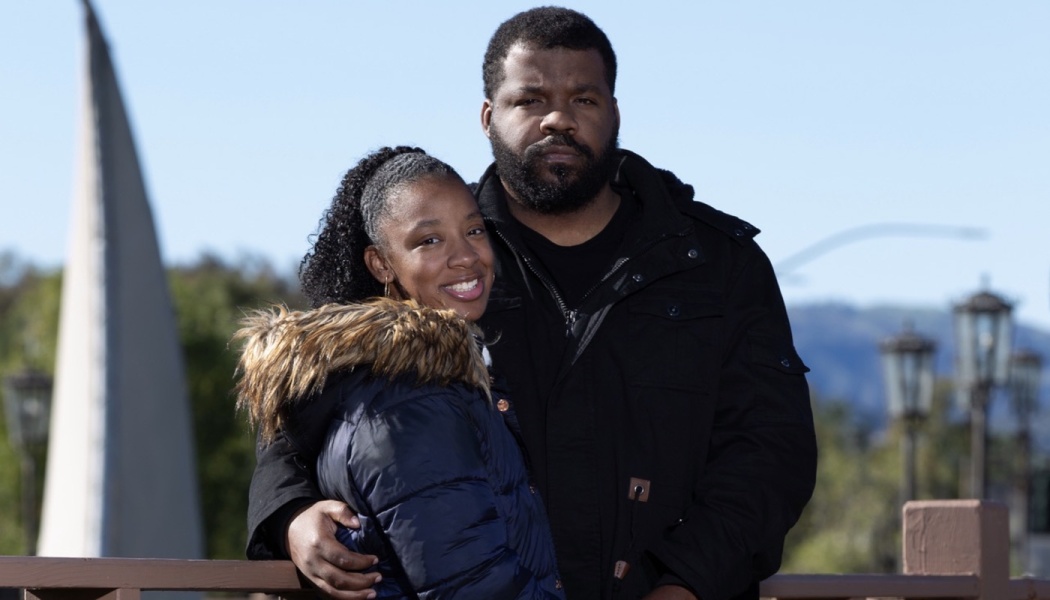 A Black couple posing outdoors in winter coats, with the man's arm around the smiling woman.