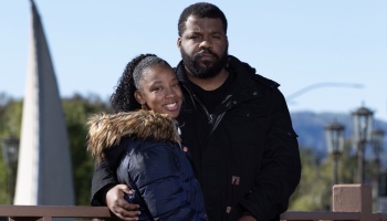 A Black couple posing outdoors in winter coats, with the man's arm around the smiling woman.