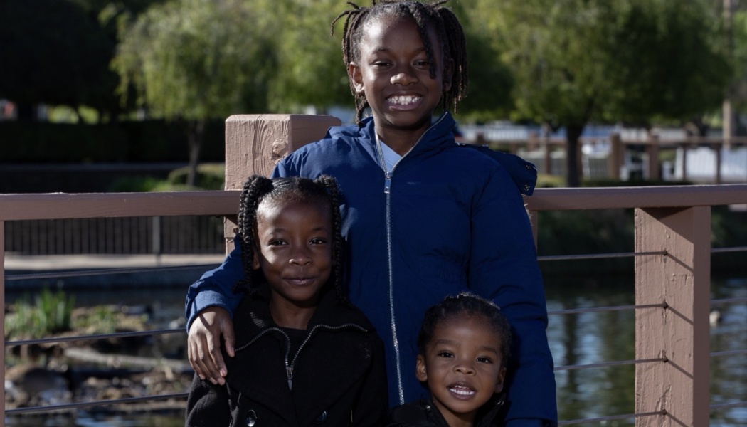 Three smiling young Black children standing together outdoors near a wooden park railing.