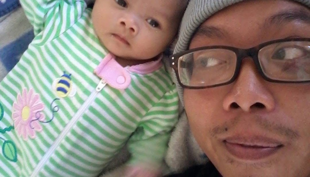 Close-up selfie of a man wearing glasses and a beanie next to a baby in a green striped onesie.
