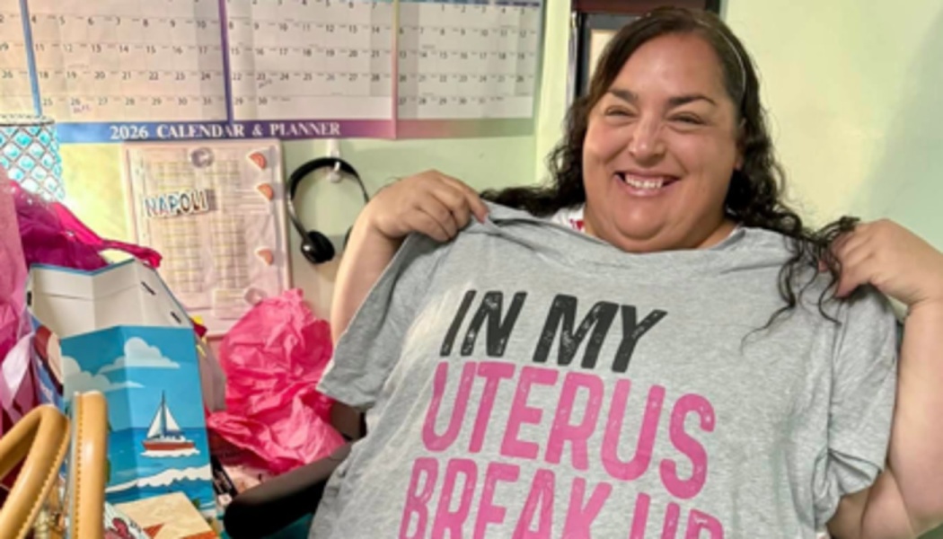 Smiling woman holding up a grey t-shirt that reads "IN MY UTERUS BREAK UP" in pink and black lettering.