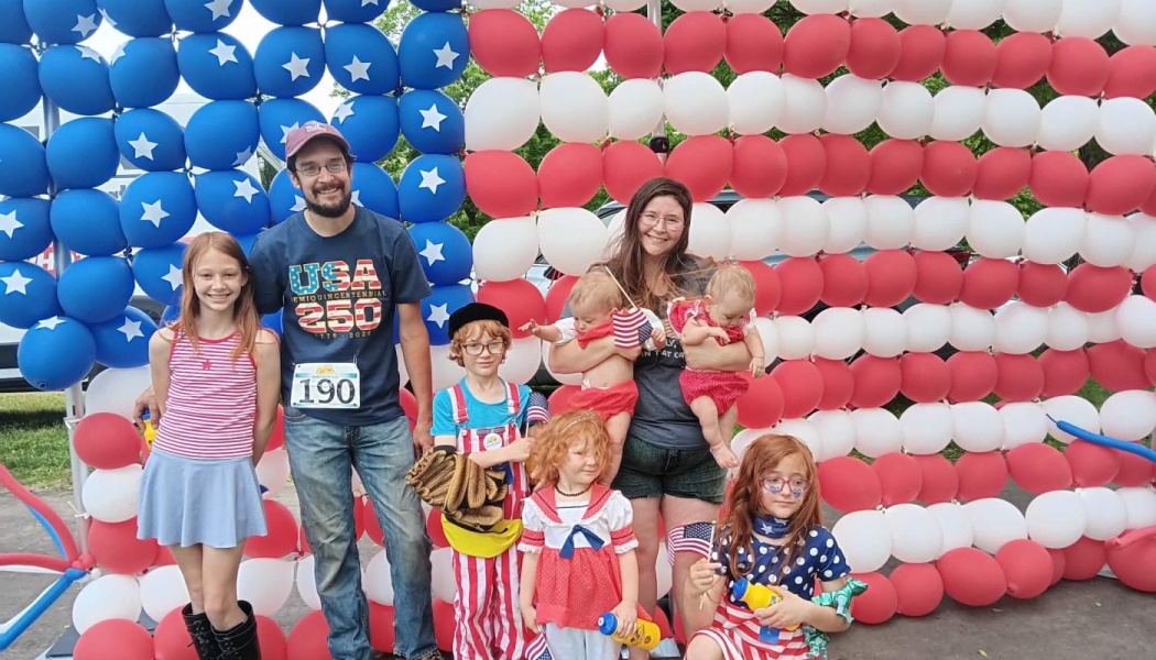 A smiling family poses together in front of a giant American flag made of red, white, and blue balloons.