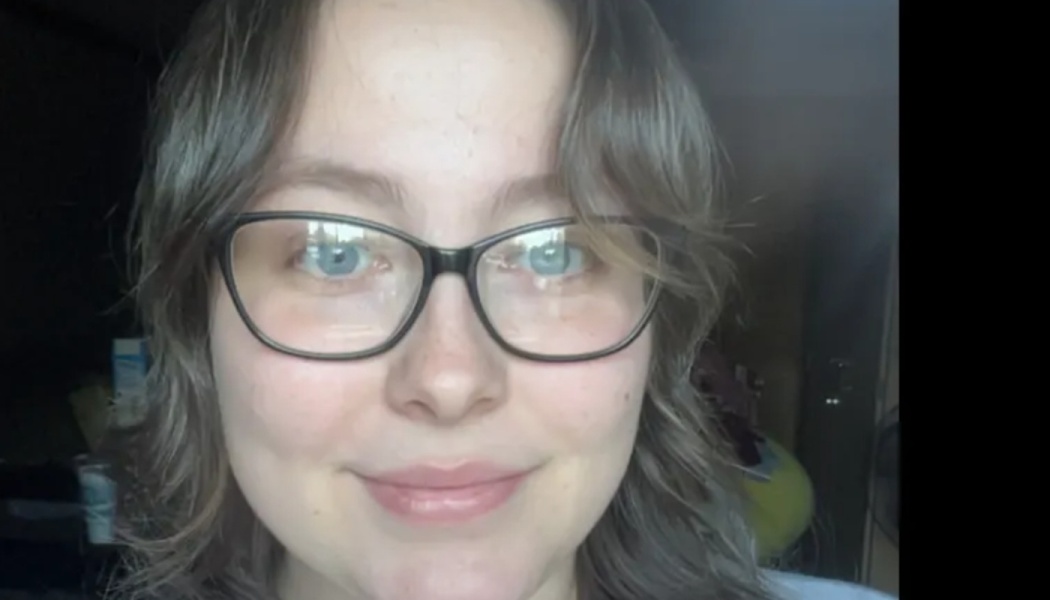 Close-up portrait of a young woman with blue eyes, wavy brown hair, and black glasses smiling gently at the camera.
