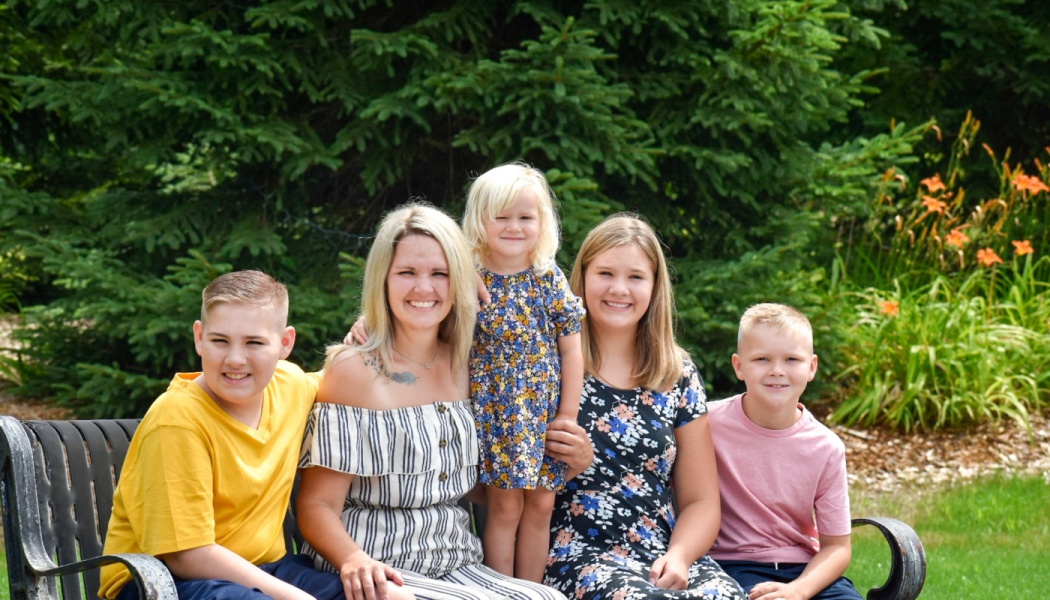 A smiling mother sits on a park bench with her four children in front of lush green foliage.
