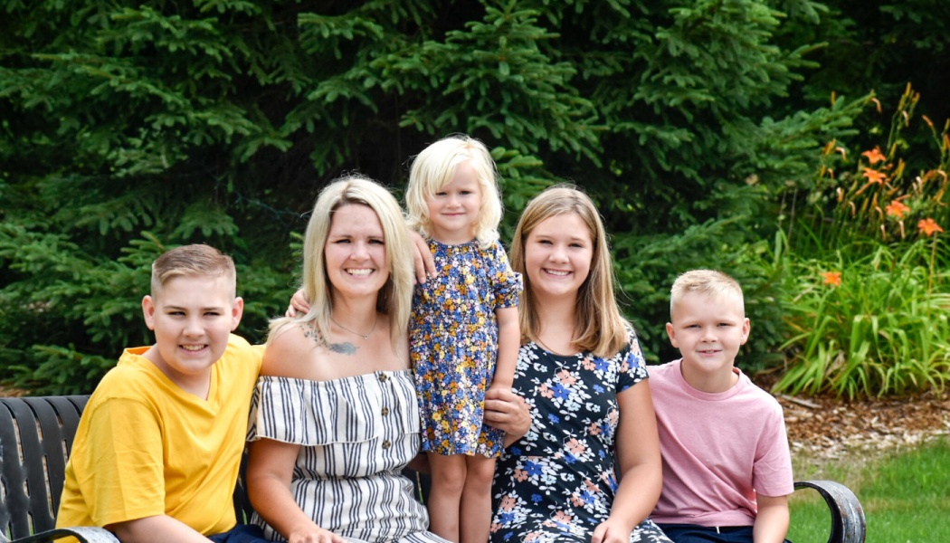 A mother and her four smiling children sit together on a bench outdoors in front of lush evergreen trees.