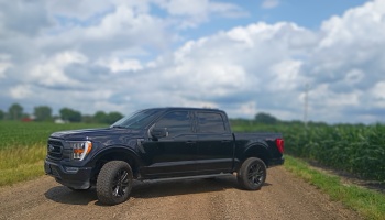 Black Ford pickup truck parked on a dirt country road beside green farm fields under a cloudy sky.